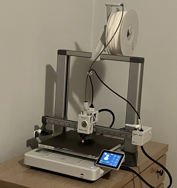 Bambu Lab A1 3D printer, in the process of constructing a 15mm white rook. The printer consists of a rectangular base; a square 256mm by 256mm build plate, which moves back and forth to constitute the Y-axis; a vertical frame straddling the base, supporting a gantry that moves up and down to constitute the Z-axis; and a rectangular tool head that moves left and right on the gantry, constituting the X-axis.  A 1kg spool of filament is mounted above the Z-axis frame, and a small touchscreen beside the build plate displays the model being printed.