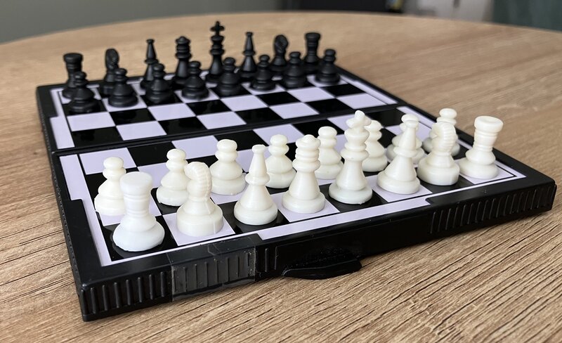 Three-quarter view of a pocket-sized magnetic chess set in starting position, from White's queenside perspective. The set features 14mm squares and a 23mm king height.
