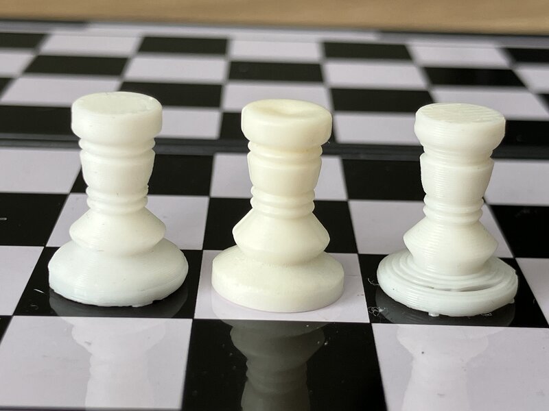 Close-up view of three 15mm white rooks, arranged side-by-side on the magnetic chessboard. The center rook is the injection-molded original, while the other two are 3D-printed. The left rook looks nearly identical to the original, while the right rook has several imperfections; most obviously a large hole in the base where the printer was unable to bridge across the gap where the magnet belongs.